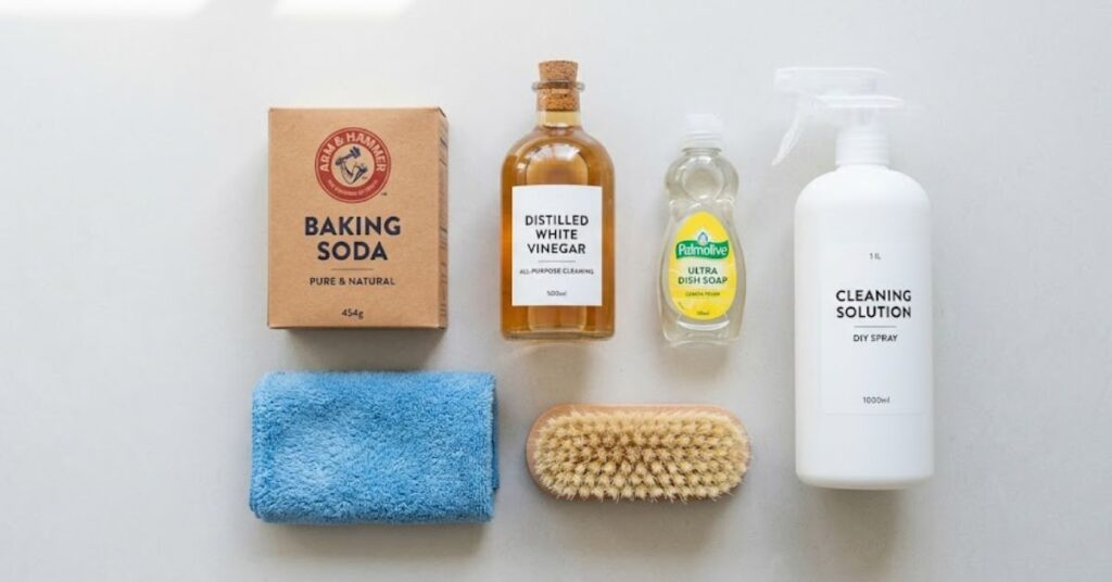 A flat lay of carpet cleaning supplies, including baking soda, distilled white vinegar, dish soap, a spray bottle, a blue microfiber cloth, and a soft brush, arranged on a clean, light-colored surface.