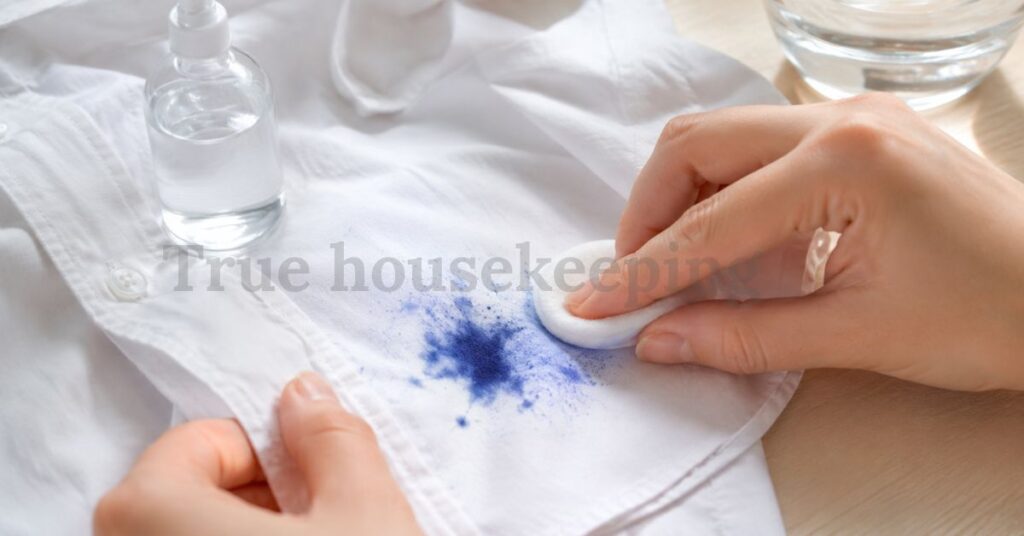 Person gently removing a blue ink stain from a white shirt using a cotton pad soaked in cleaning solution.
